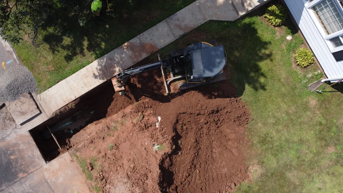 Overhead view of an excavator beside an open trench along a residential walkway